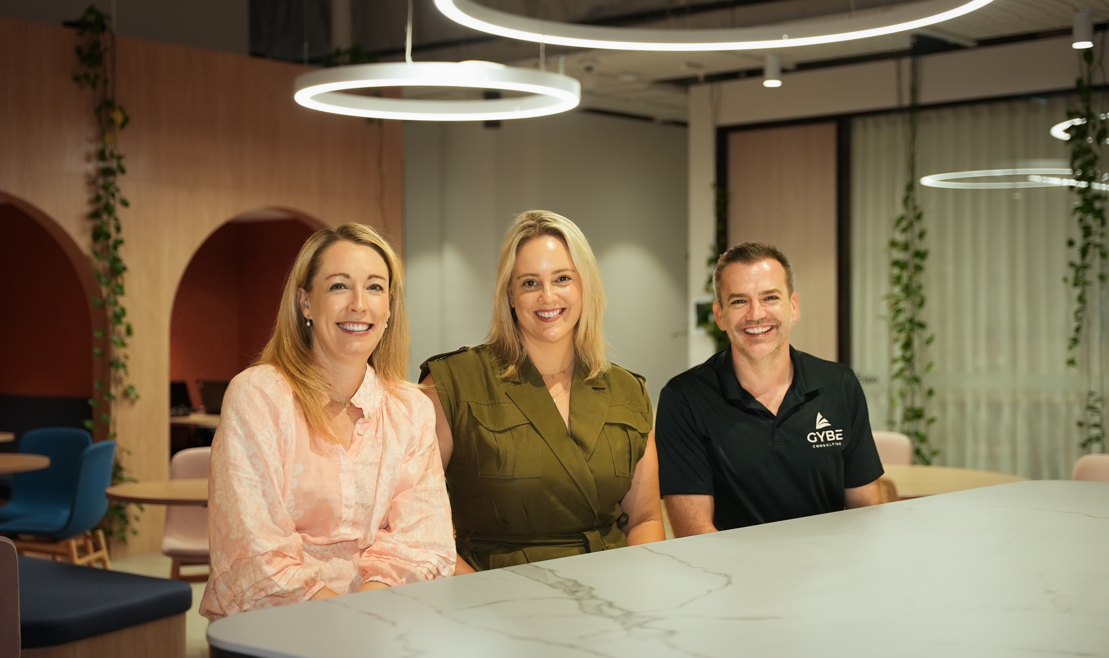 Three people smiling and sitting at a marble table in a modern office with circular lights and plants.