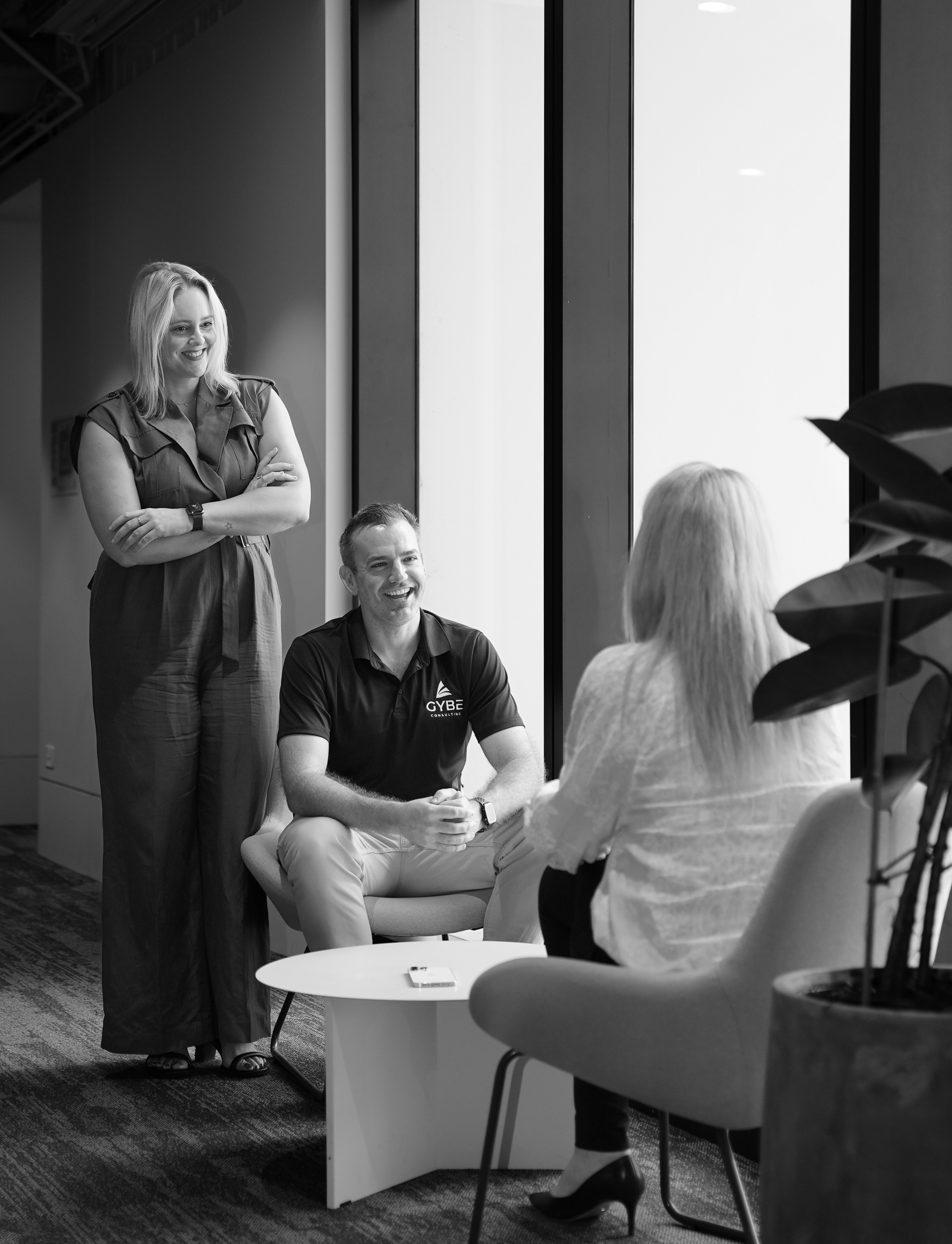 Black and white photo of three people in a relaxed office setting, two sitting and one standing near a window.