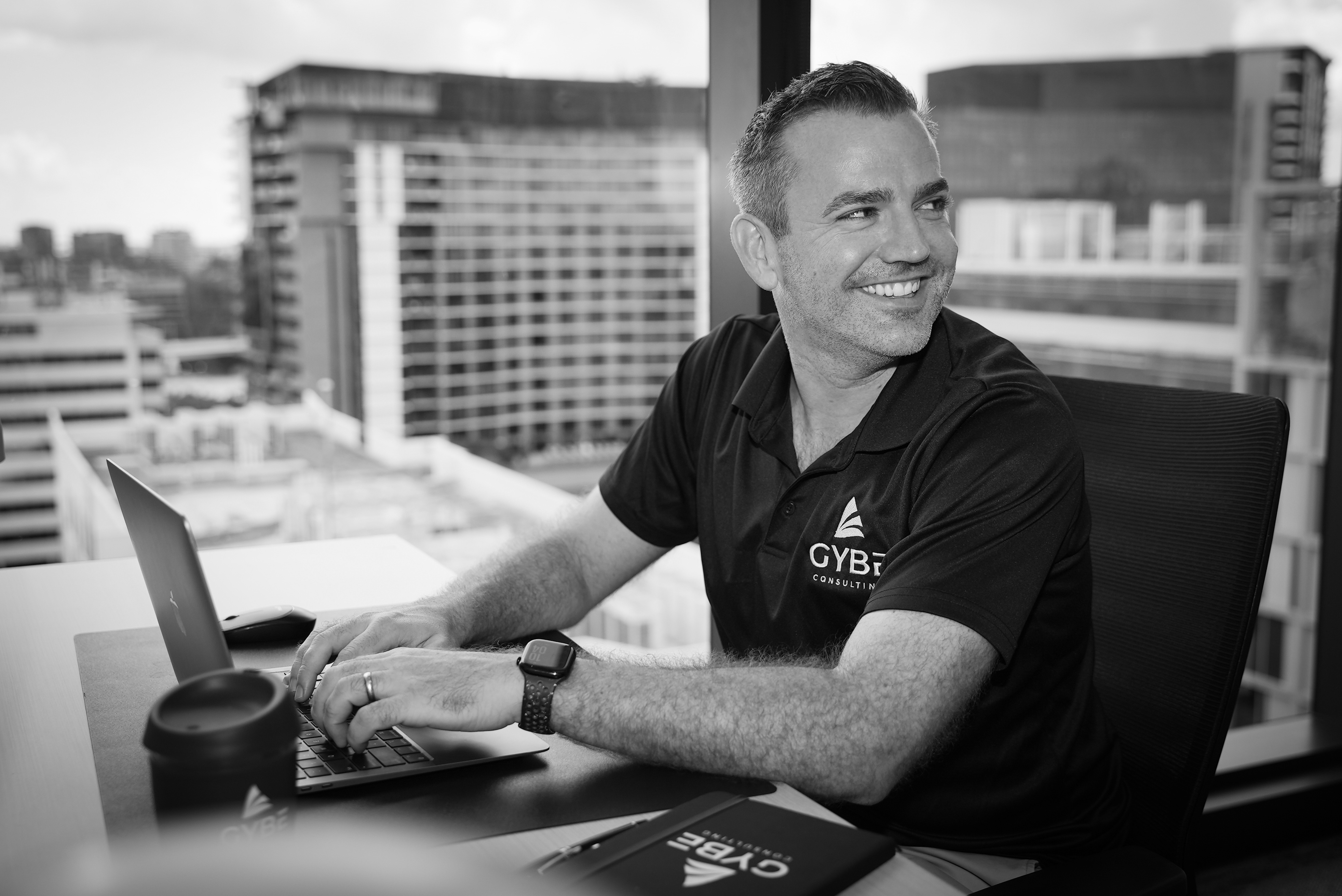 Smiling man in a black GYBE Consulting polo shirt typing on a laptop at a desk with city buildings visible through a window behind him.