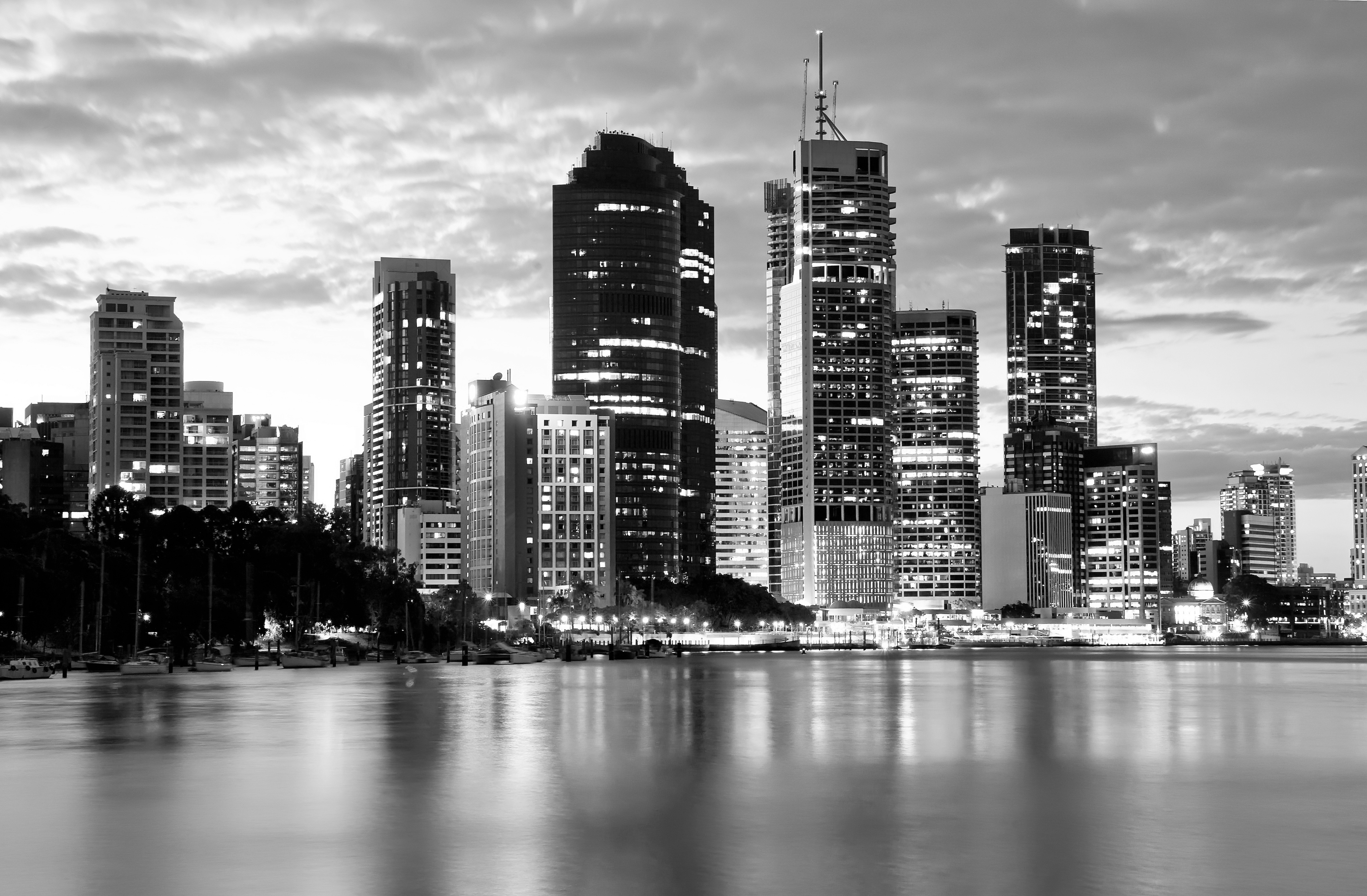 Black and white city skyline with illuminated skyscrapers reflecting on a body of water at dusk.